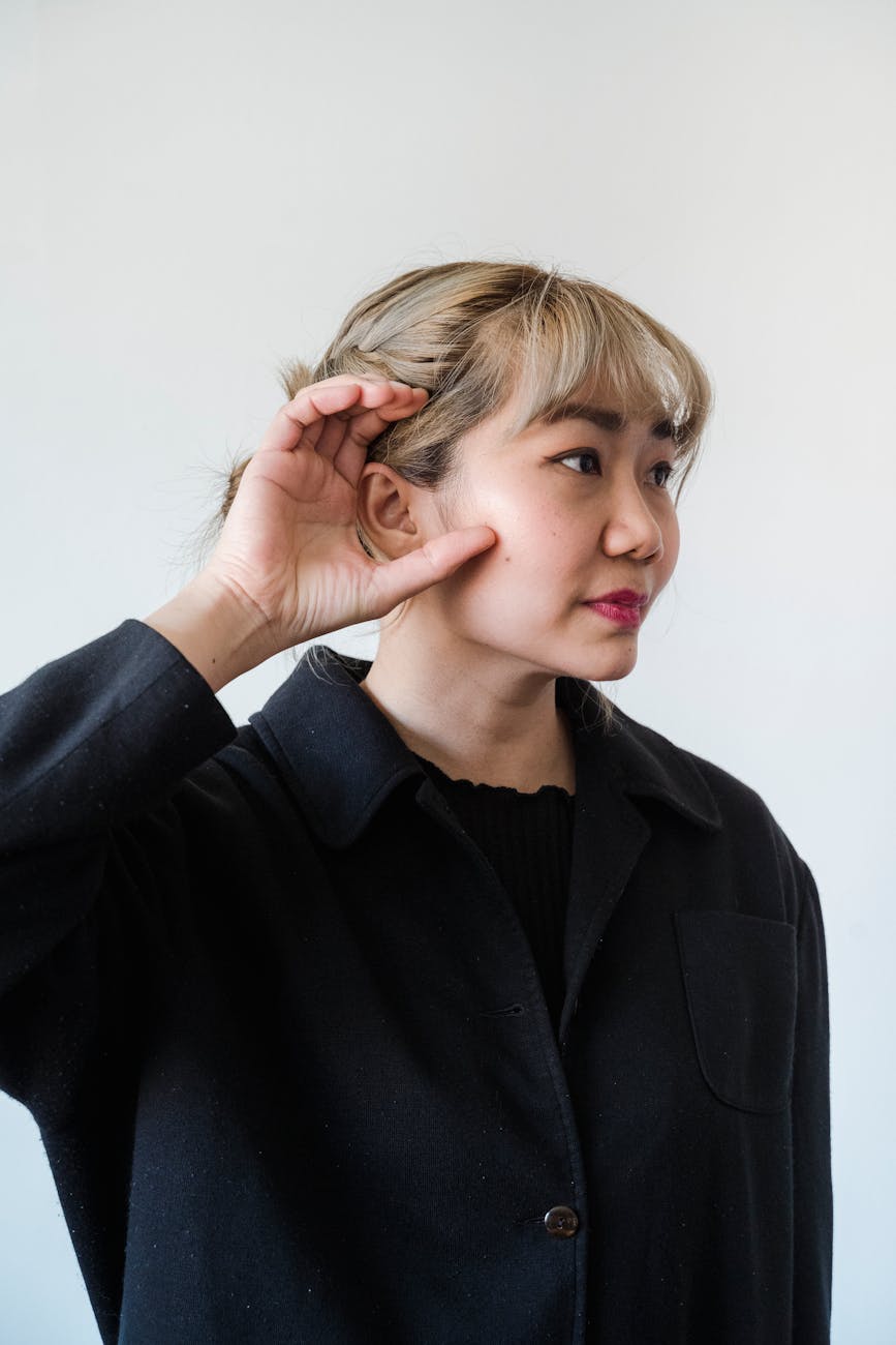 A woman with fair hair wearing a black coat holds her hand next to her ear, as if listening closely, against a plain light background.