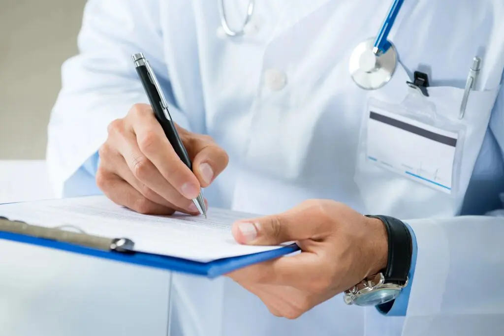 A doctor in a white coat and stethoscope writes on a clipboard with a pen, with an identification badge clipped to the coat pocket.