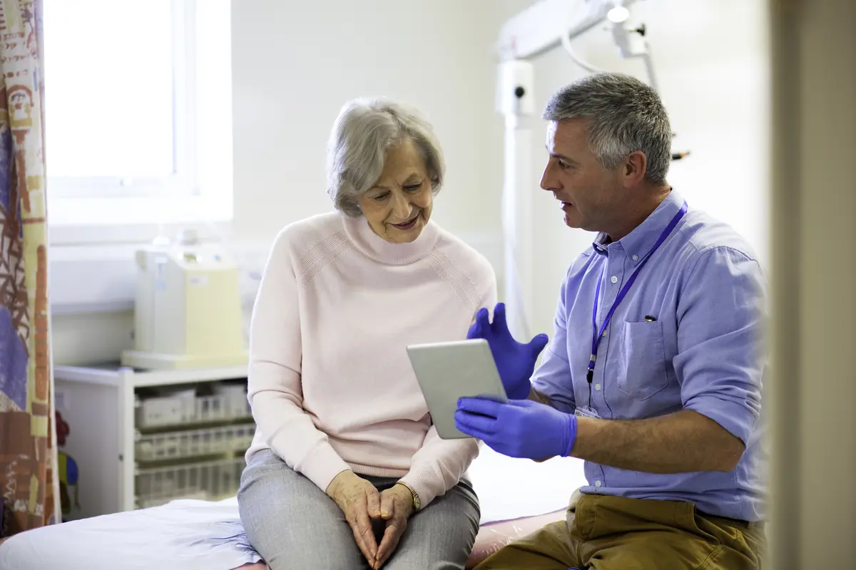 A healthcare professional wearing gloves shows a digital tablet to an older woman sitting on an examination table in a medical surgery.