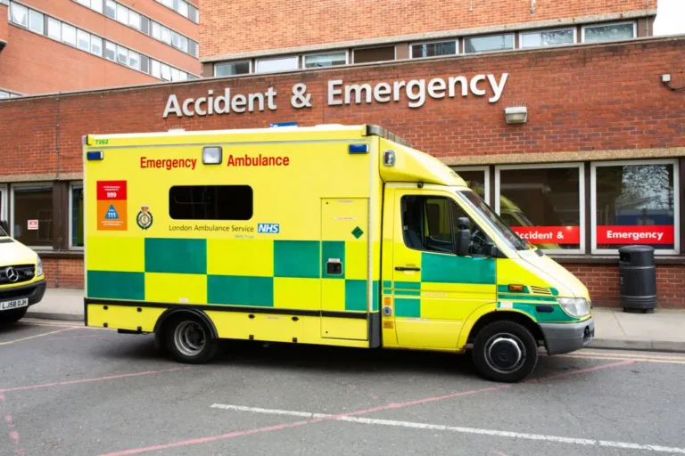 A yellow and green London Ambulance Service vehicle parked outside an Accident & Emergency entrance at a hospital.