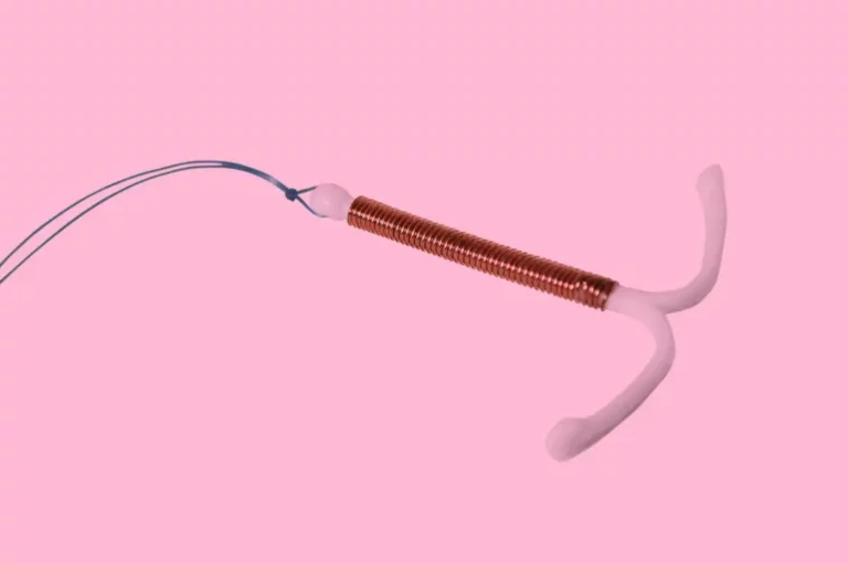A copper intrauterine device (IUD) with plastic arms and string, shown on a pink background.