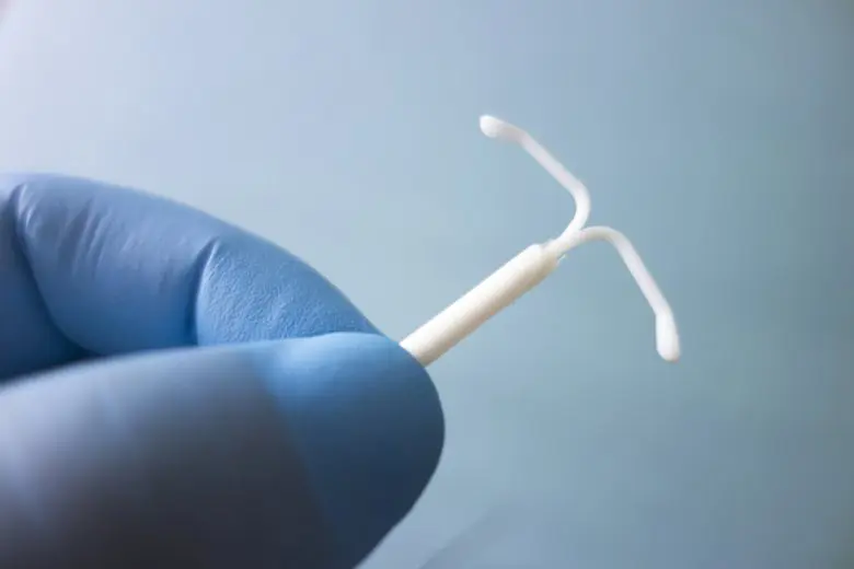 A gloved hand holds a white intrauterine device (IUD) against a plain blue background.
