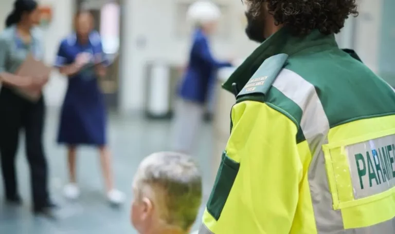 A paramedic in uniform stands beside a seated person in a hospital corridor as medical staff converse in the background.