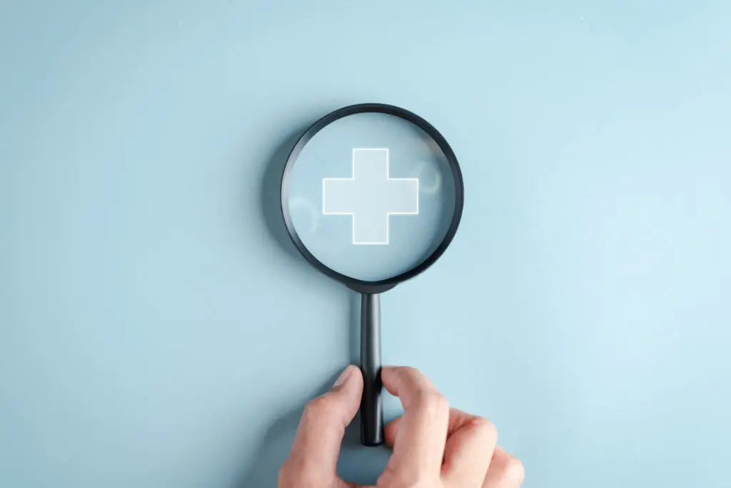 A hand holds a magnifying glass over a white medical cross symbol on a light blue background.