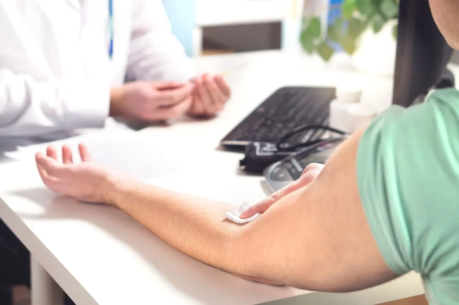 A personโs arm with a plaster at the elbow rests on a desk opposite a healthcare professional, with a computer and medical equipment in the background.