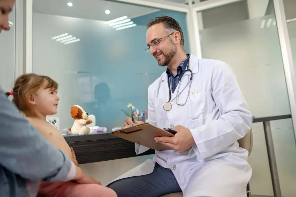 A doctor in a white coat sits with a clipboard, smiling and talking to a young girl and her parent in a surgery.