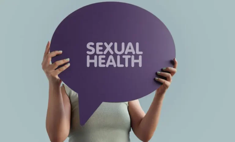 A person holds a large purple speech bubble sign with the words "SEXUAL HEALTH" printed in white.