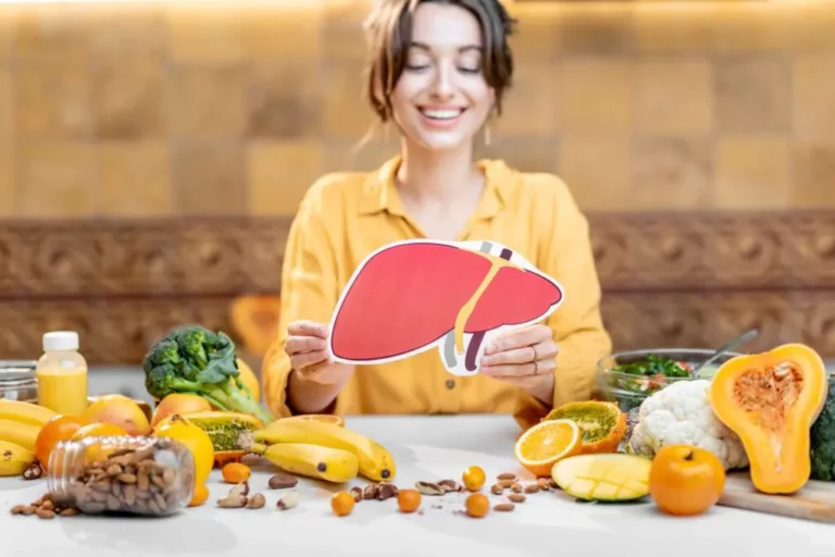 A woman holding a paper illustration of a liver sits at a table with various fruits, vegetables, nuts, and seeds in front of her.
