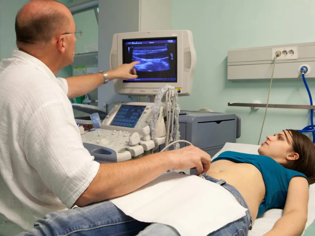 A doctor performs an abdominal ultrasound on a woman lying on an examination table, while pointing at the monitor displaying the scan.