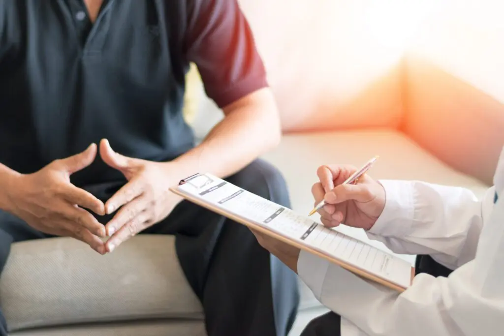 A person sits with hands clasped whilst another person in a white coat writes on a clipboard during a consultation.