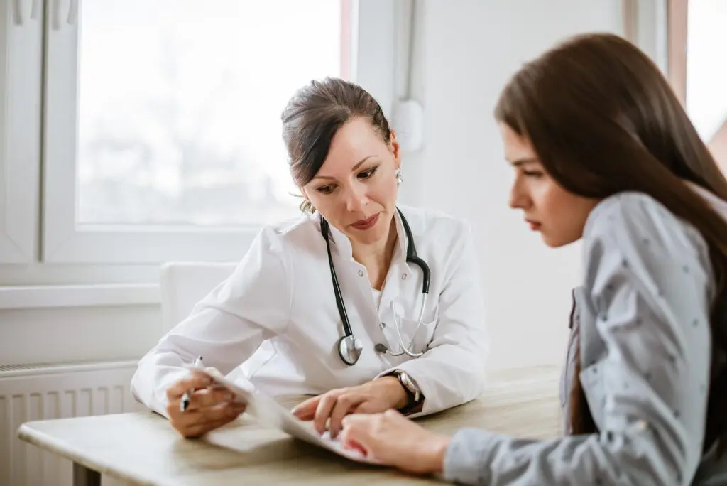 A doctor points to a document while discussing it with a patient at a desk in a medical surgery.