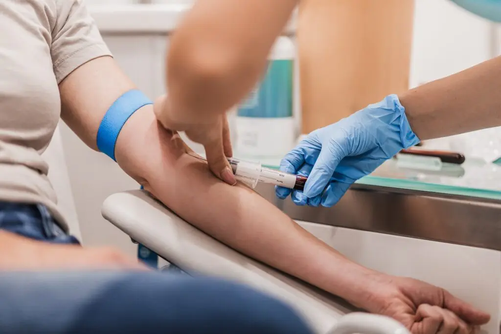 A healthcare professional wearing gloves takes blood from a person's arm using a syringe. The person has a blue tourniquet on their upper arm.