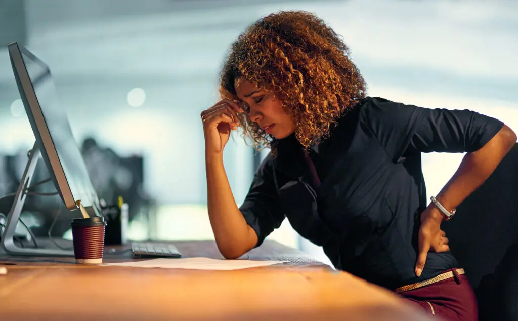 A woman sitting at a desk grimaces in pain, holding her lower back with one hand whilst looking at paperwork beside a computer monitor.