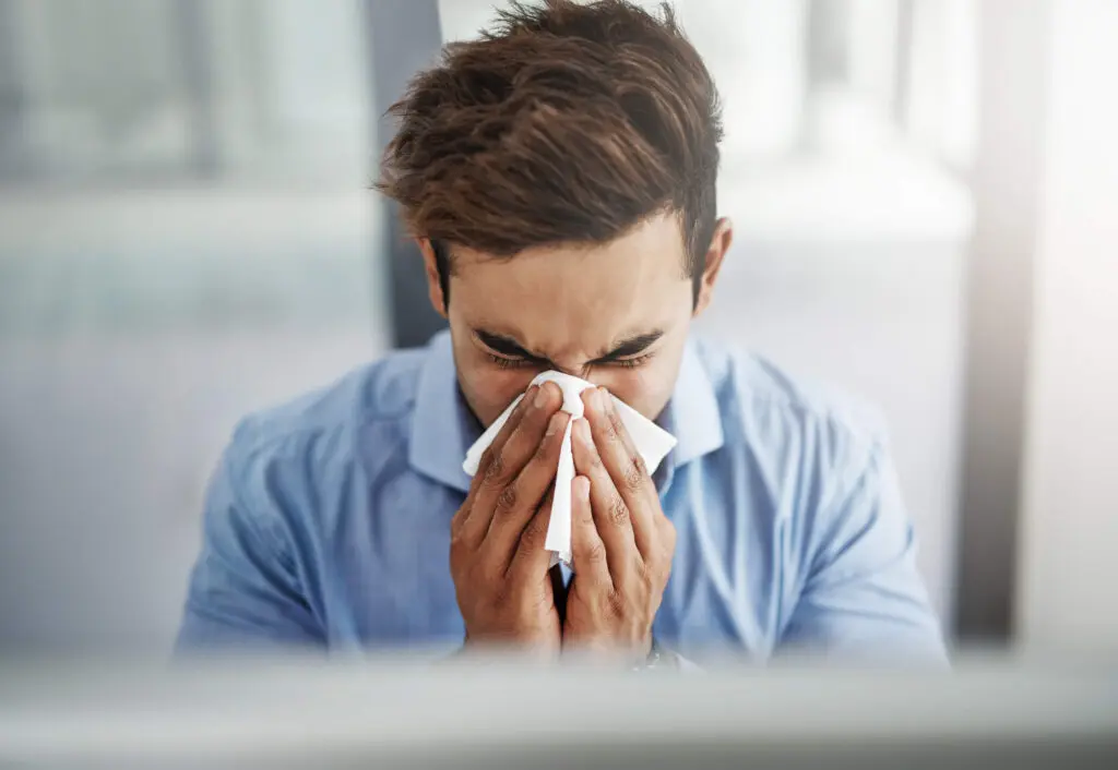 A man in a blue shirt is sitting indoors, holding a tissue to his nose and sneezing or blowing his nose.