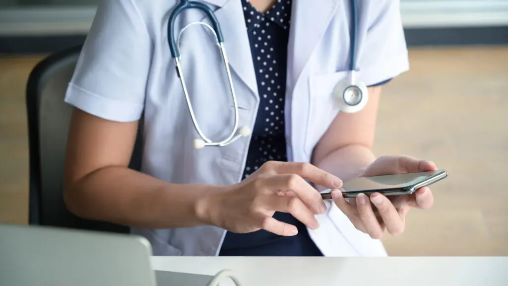 A person wearing a white medical coat and stethoscope uses a mobile phone while seated at a desk.