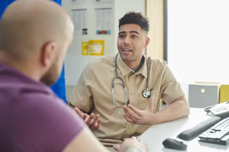 A doctor with a stethoscope around his neck talks to a patient across a desk in a surgery.