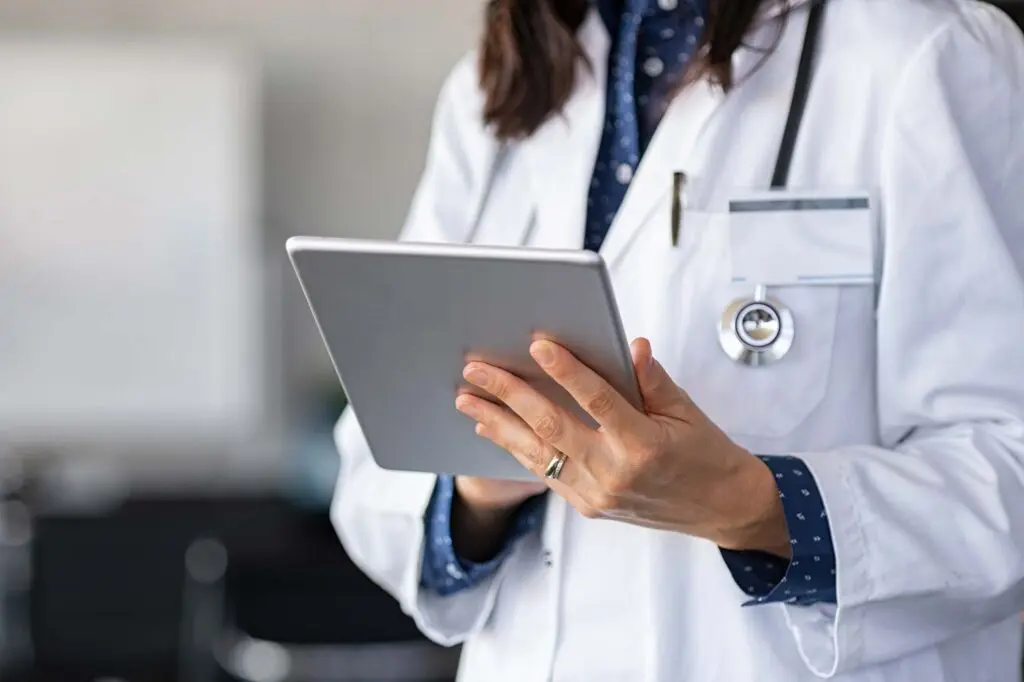 A person wearing a white lab coat and stethoscope is holding and using a tablet device.