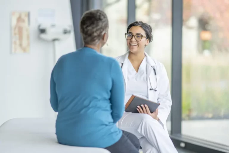 A doctor in a white coat sits and smiles whilst talking with a patient in blue clothing during a medical consultation in a consulting room.