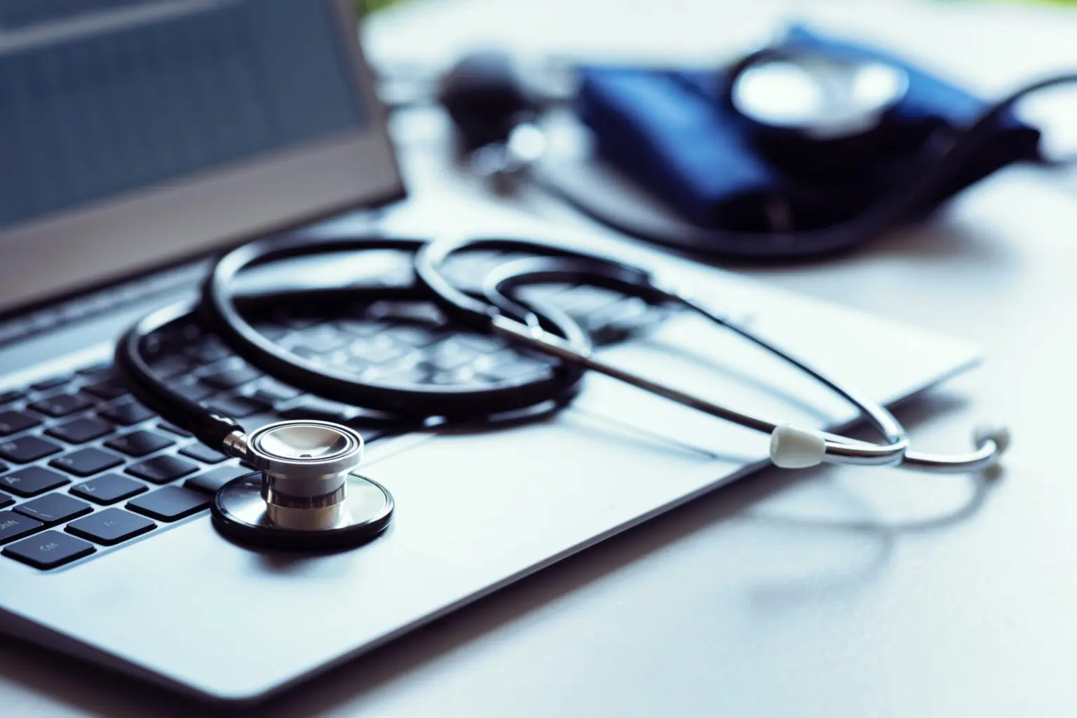 A stethoscope rests on a laptop keyboard, with a blood pressure cuff in the background on a desk.