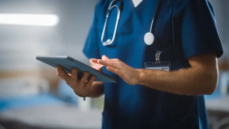 A medical professional in blue scrubs with a stethoscope uses a digital tablet in a clinical setting.