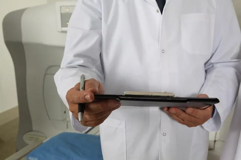 A person in a white lab coat holds a clipboard and pen, standing in a clinical or medical setting next to medical equipment.