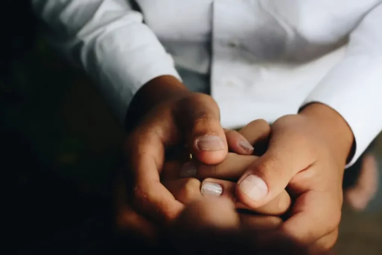 Close-up of an adult gently holding a child's hand, both wearing white clothing, with a blurred dark background.