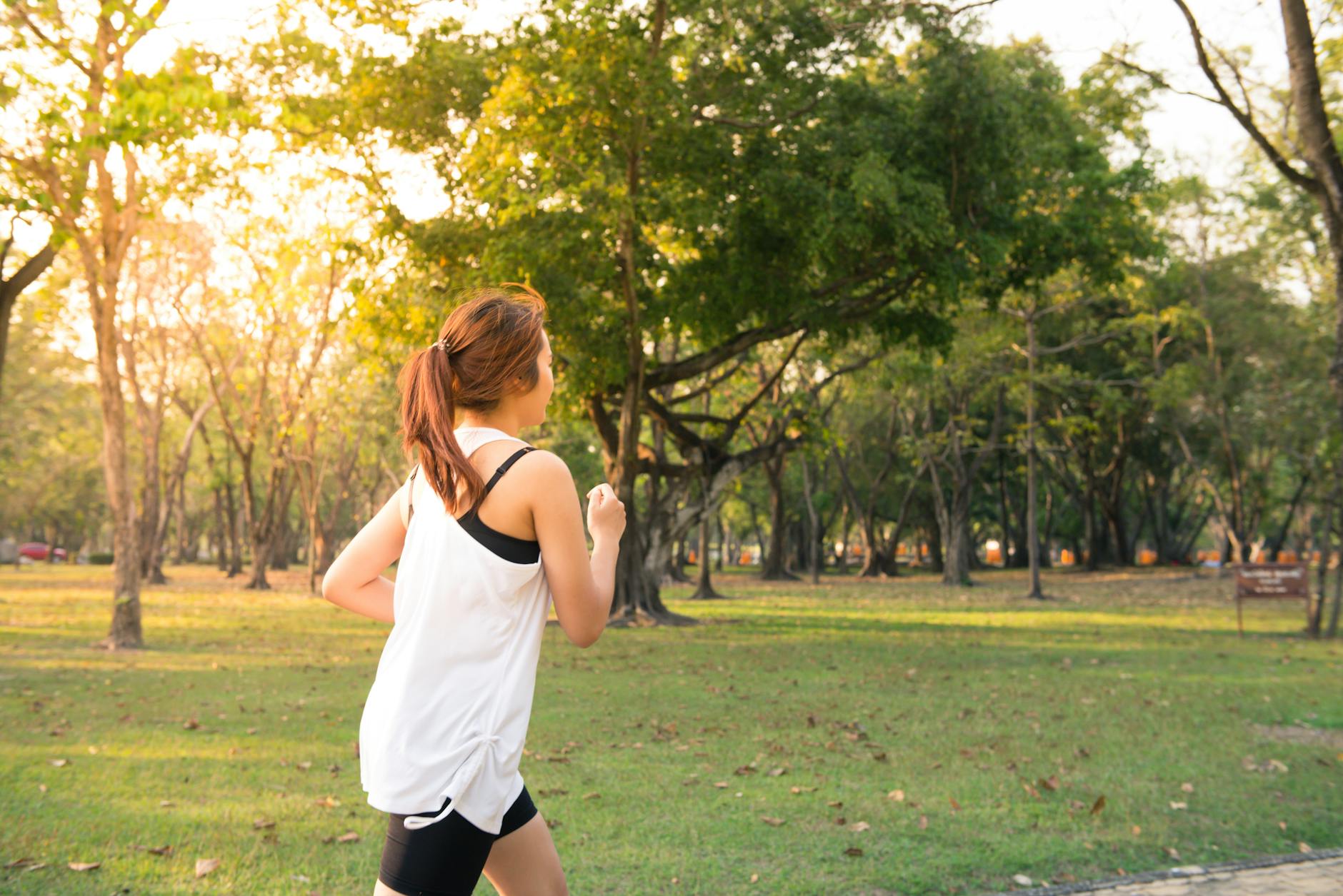 A woman in sportswear jogs through a grassy park area with trees and sunlight in the background.