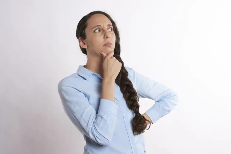 A woman with a long plait wearing a light blue shirt stands with one hand on her chin and looks upwards, appearing thoughtful, against a plain white background.