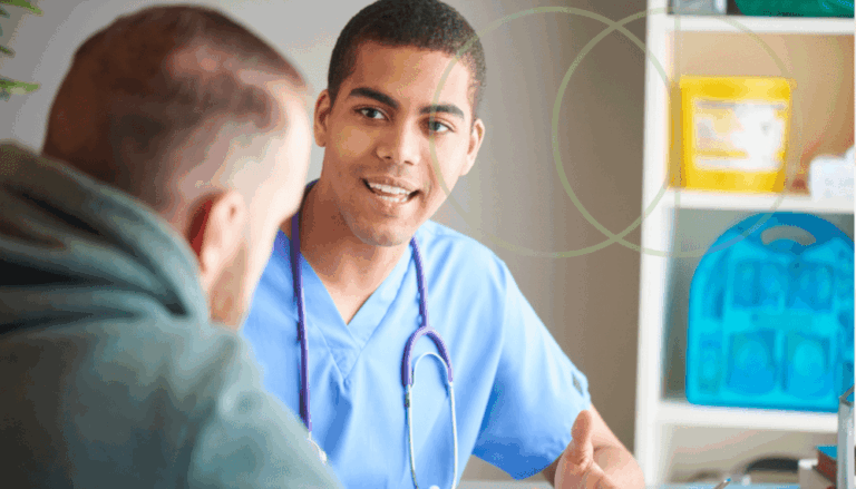 A healthcare professional in blue scrubs and a stethoscope talks with a patient in an office setting. Medical supplies are visible on shelves in the background.