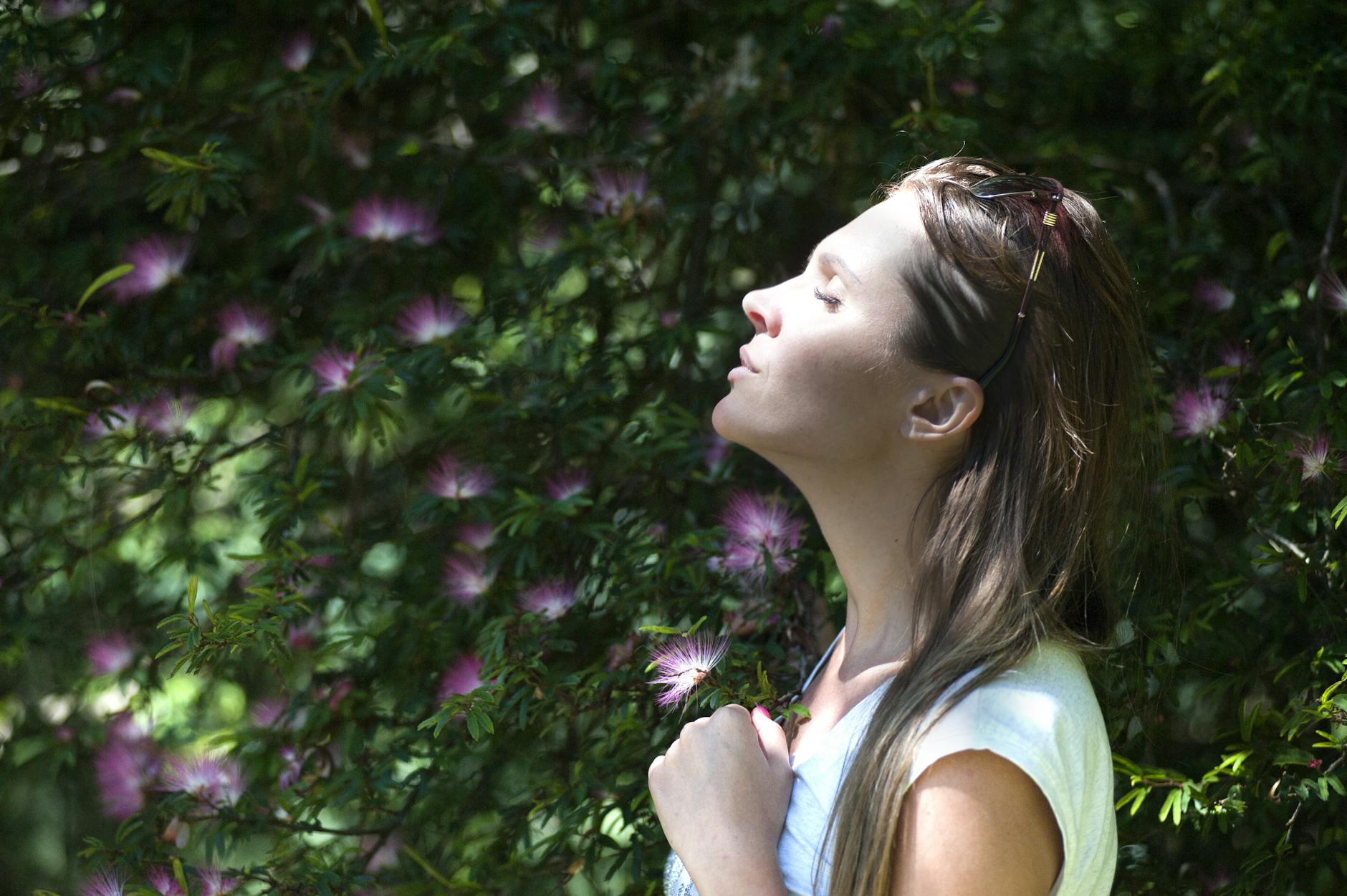 A woman stands outdoors with her eyes closed, facing the sunlight, surrounded by green foliage and pink flowers.