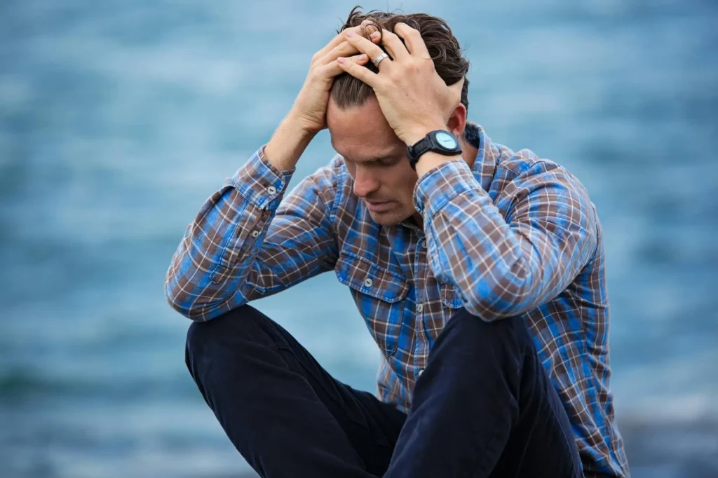 A man in a checked shirt sits with his elbows on his knees, holding his head in his hands, appearing stressed, with water in the background.