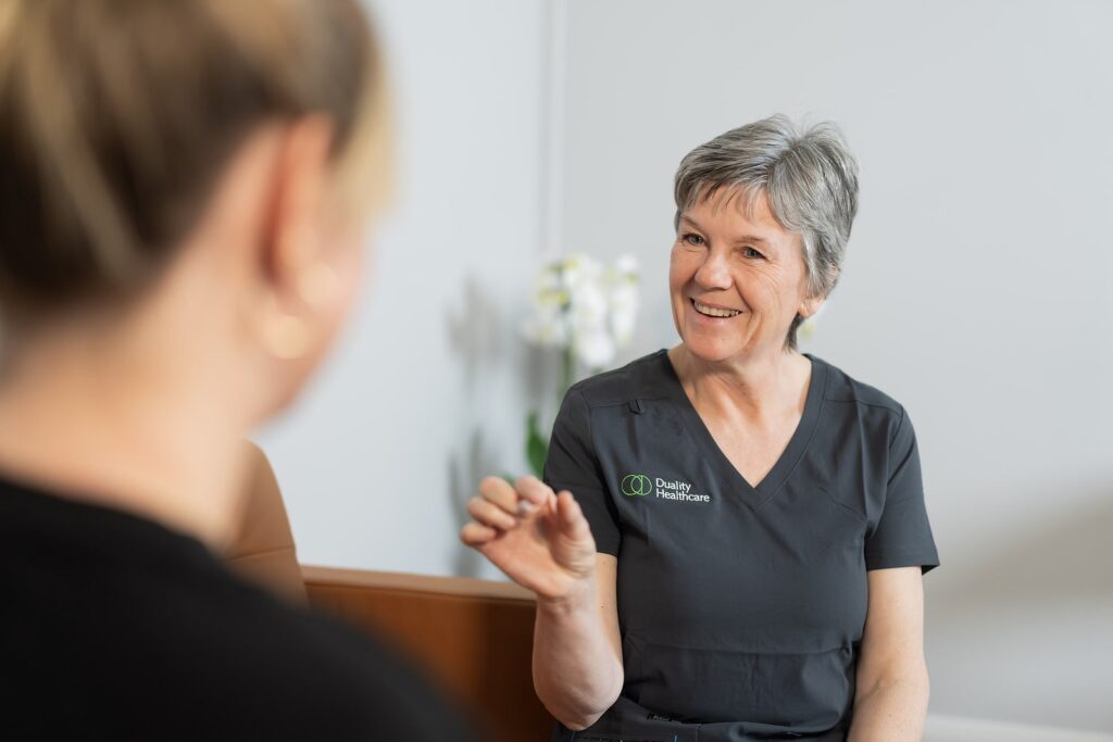 A healthcare professional in scrubs smiles and gestures whilst speaking with a seated person in an indoor setting.