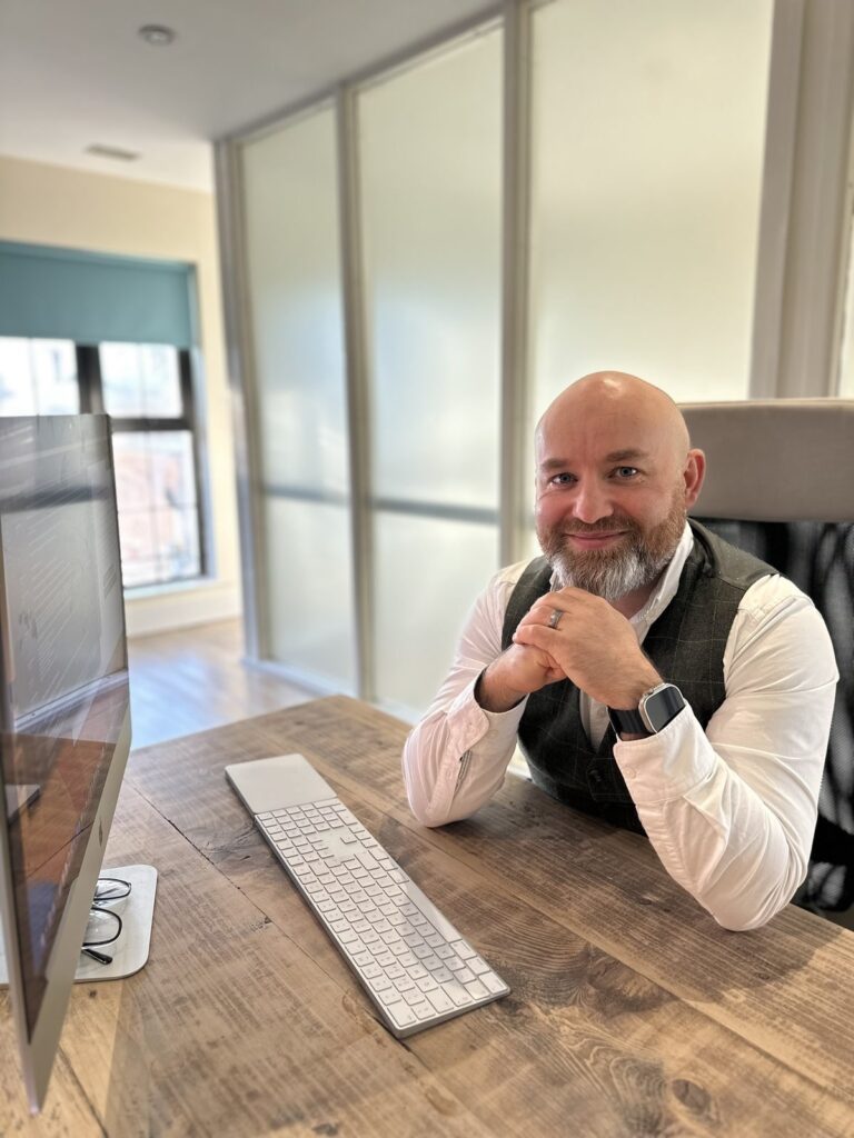 A man with a beard sits at a desk with a computer, keyboard, and mouse in a modern, well-lit office space.