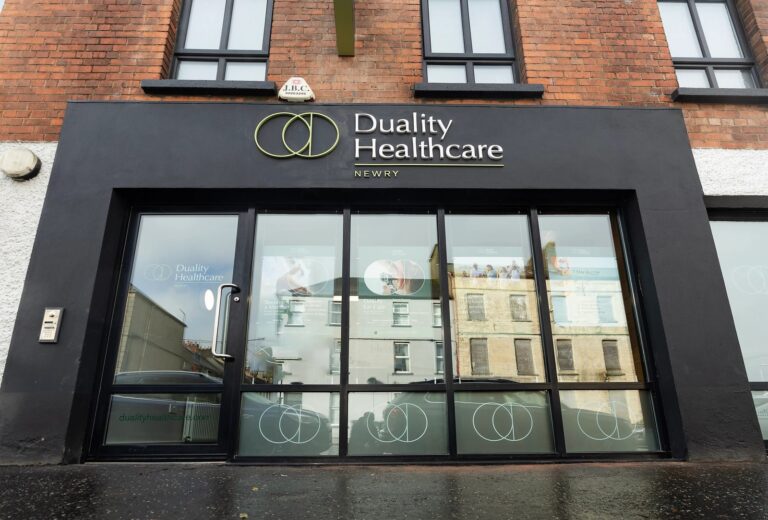 The entrance of Duality Healthcare Newry, featuring glass doors with logo decals and the clinic’s name displayed above in white lettering on a black sign.