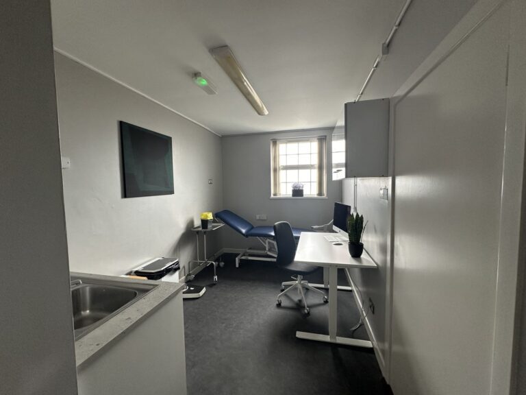 A small, clean medical examination room with a desk, computer, examination couch, sink, and window providing natural light.
