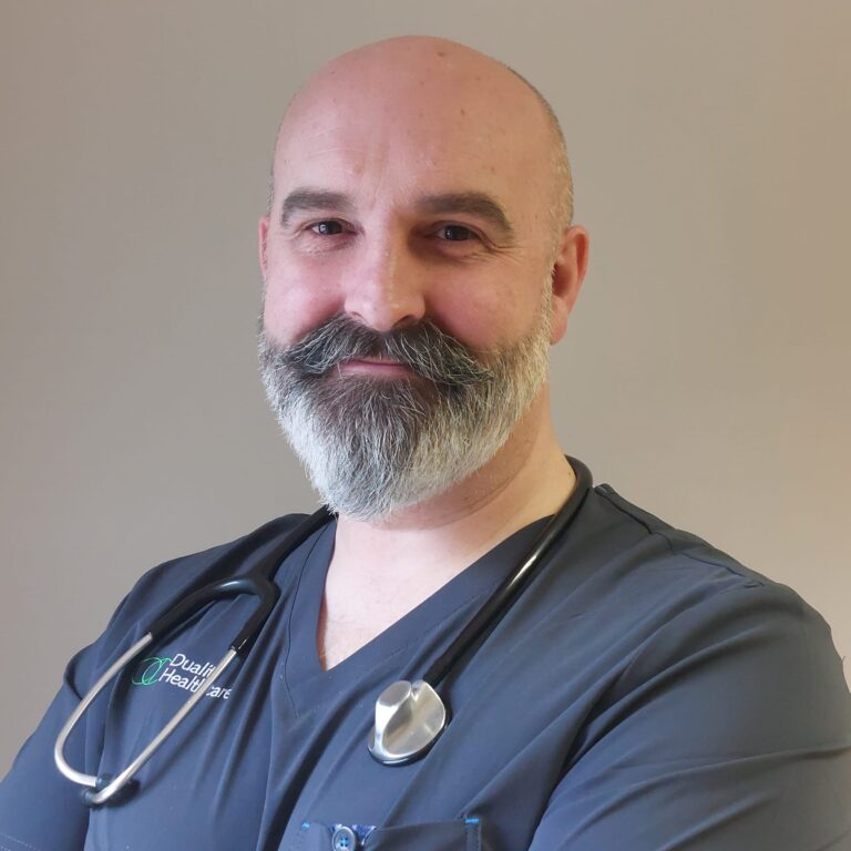 A middle-aged man with a beard and moustache, wearing medical scrubs and a stethoscope, stands with arms crossed and smiles at the camera.