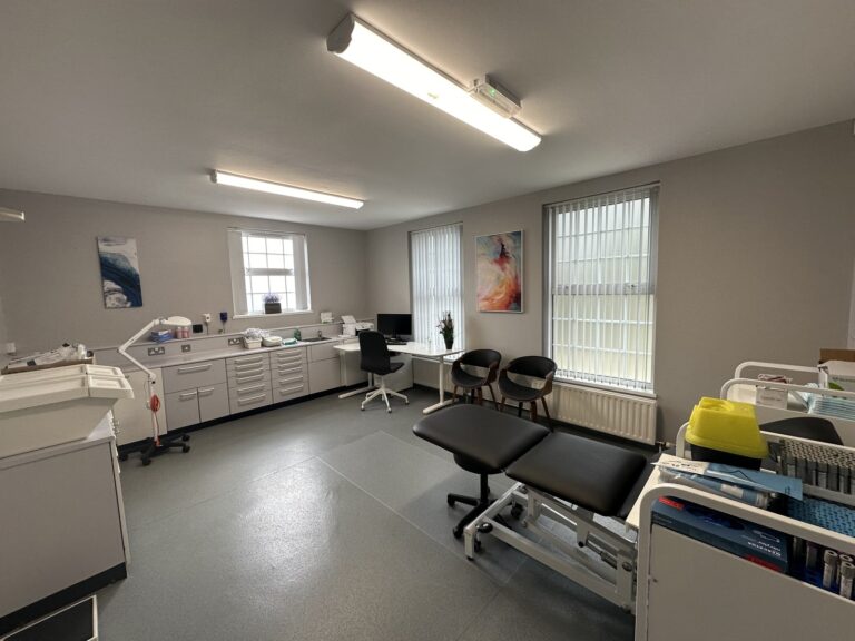 A medical examination room with an examination couch, two chairs, medical equipment, cupboards, a desk with a computer, and bright overhead lights.