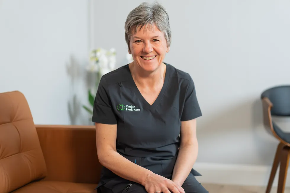 A woman in dark scrubs with a "Quality Healthcare" logo sits on a brown sofa, smiling in a modern, well-lit room.
