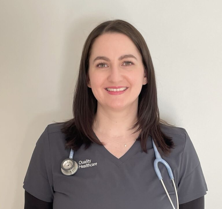 A woman in dark scrubs with a stethoscope around her neck stands against a plain background. Her top reads "Quality Healthcare." She is smiling and has straight, shoulder-length hair.