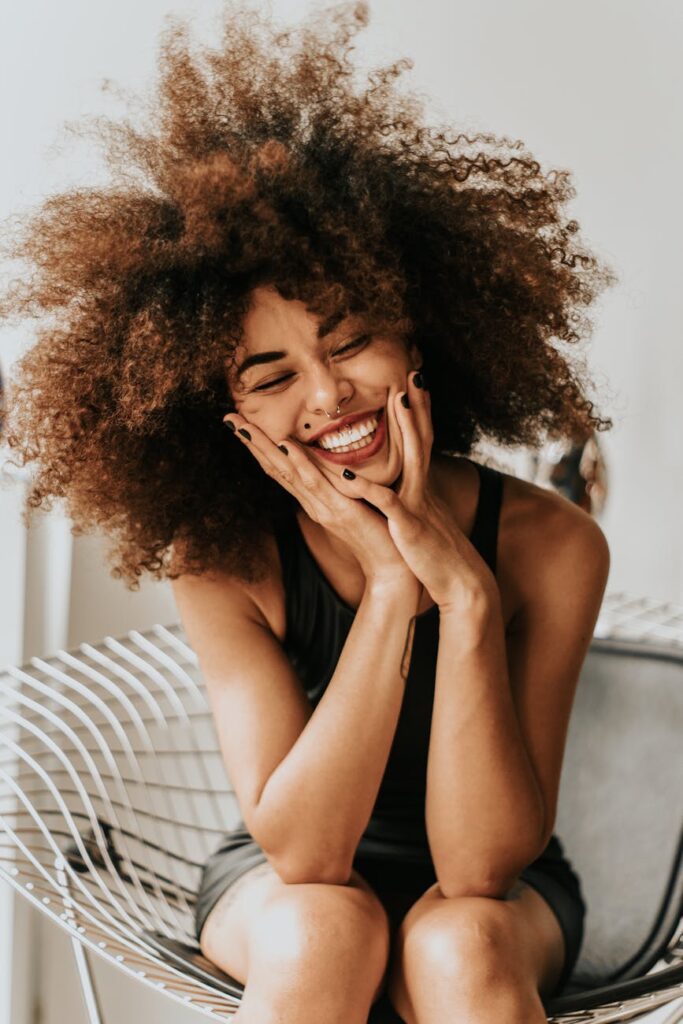 A woman with curly hair sits on a wire chair, smiling with her eyes closed and hands resting on her cheeks, wearing a sleeveless black top.