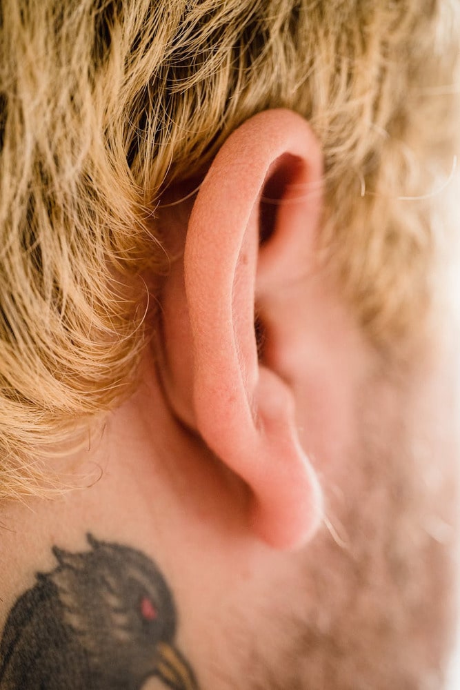 Close-up of a person's ear with blonde hair and part of a dark tattoo visible on the neck.