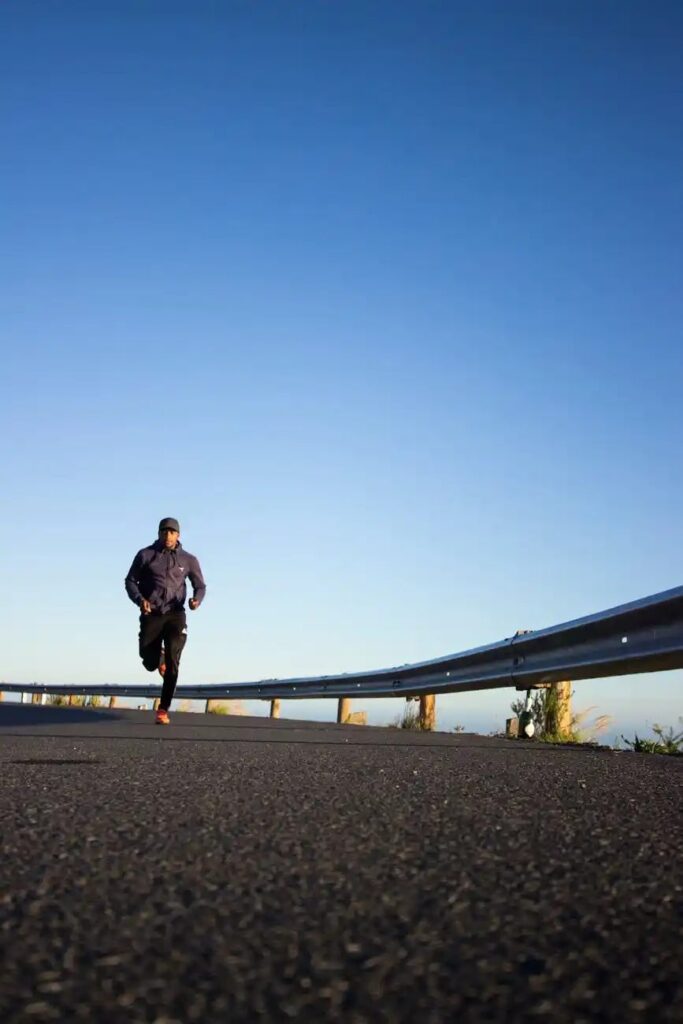 A person in sportswear runs on an empty road beside a metal crash barrier under a clear blue sky.