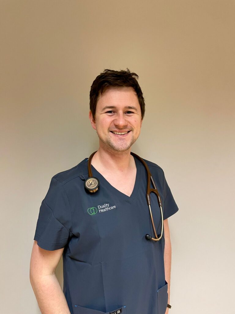 A healthcare professional wearing navy scrubs with a stethoscope around the neck stands against a plain background, smiling at the camera.