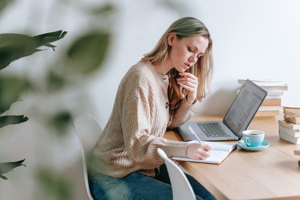 A woman sits at a desk with a laptop, writing in a notebook. A cup of coffee, books, and a plant are also on the desk.