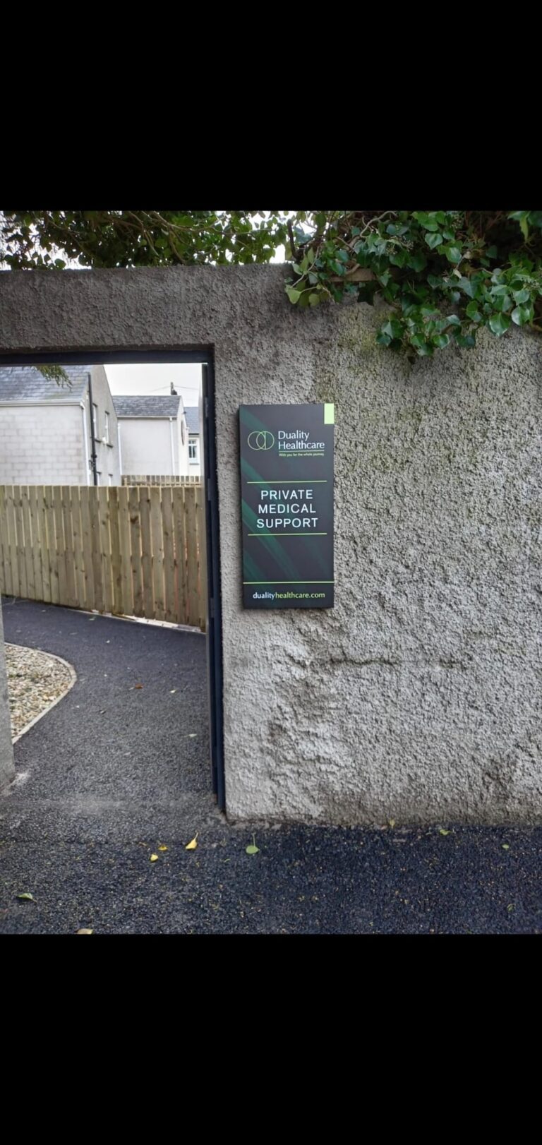 A sign on a grey wall reads “Private Medical Support” and “Duality Healthcare”, with an open gate leading to a path and a fenced area.