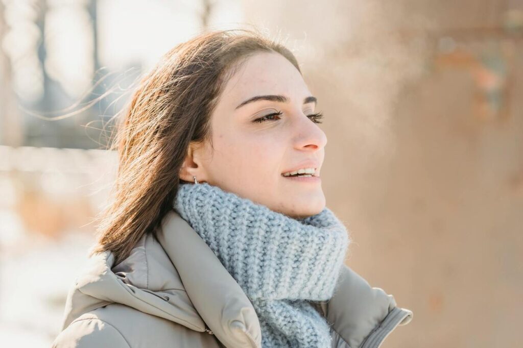 A woman in a grey coat and chunky knitted scarf stands outside on a sunny winter day, with visible breath in the cold air.