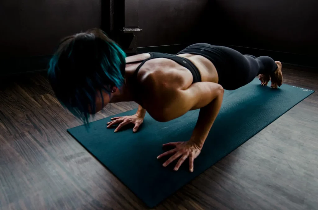 Person with short blue hair performs a chaturanga pose on a yoga mat in a dimly lit room with wooden floors.